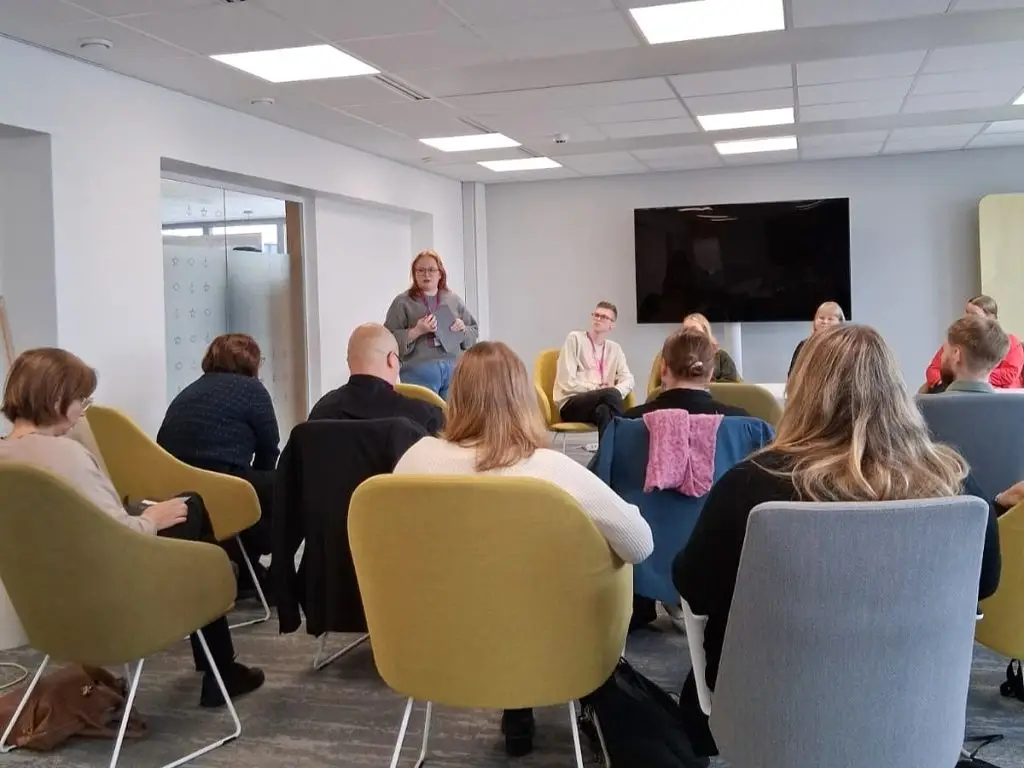 Ihmisiä kuvattuna takaapäin istumassa tuoleilla katse tilan etuosaan. Huoneen etuosassa nuoria aikuisia istumassa, sekä yksi seisomassa ja puhumassa.