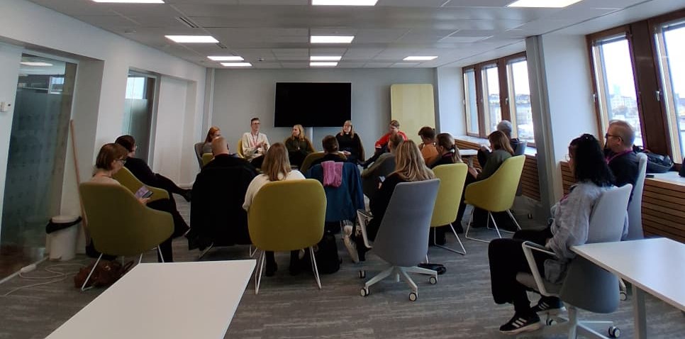 Viisi nuorta aikuista istuu tuoleilla tilan etuosassa. Heidän edessään yleisössä istuu yli kymmenen aikuista, jotka seuraavat edessä käytävää keskustelua.