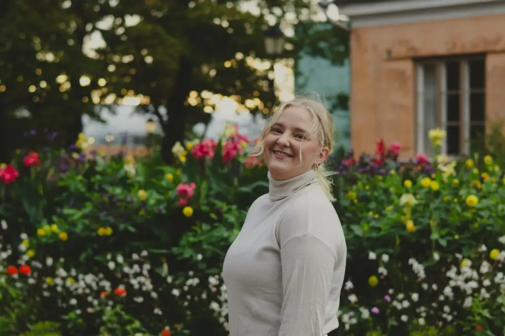 Nuori katsoo hymyillen kameraan ulkona otetussa kuvassa. Taustalla kukkia.